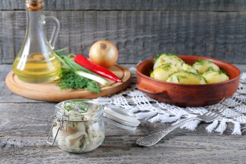 On the table is jar of salted herring near the plate with boiled potatoes and fresh greens.