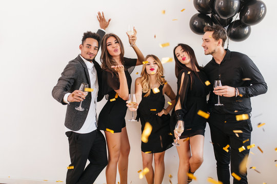 Slim european woman with tanned skin sends air kiss standing under confetti. Indoor portrait of chilling friends posing with balloons and champagne in home.