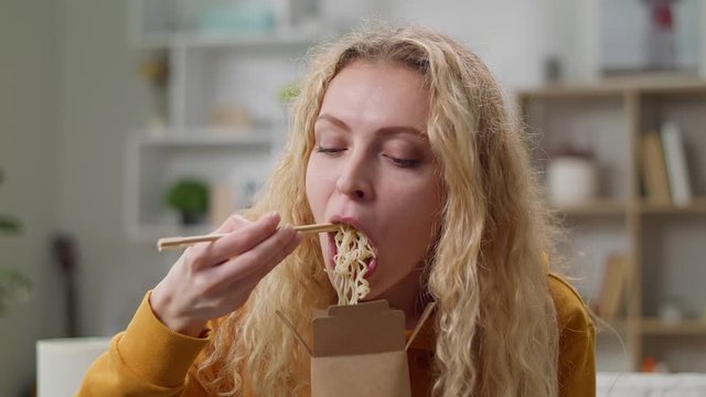 Young woman eating noodles from a box with chopsticks at home on a couch
