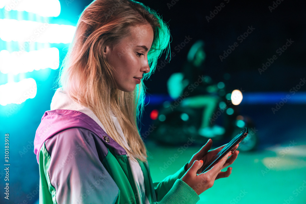 Poster Image of woman holding smartphone over multicolored neon text sign outdoors