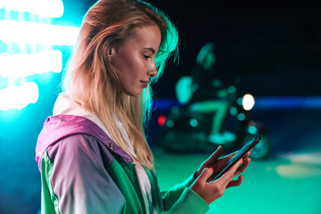 Image of woman holding smartphone over multicolored neon text sign outdoors