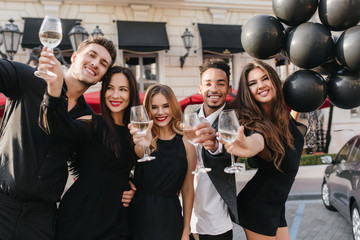 Graceful girl with long light-brown hair holding black helium balloons in front of building and laughing. Cute blonde woman raising glass of champagne while posing with african friend.