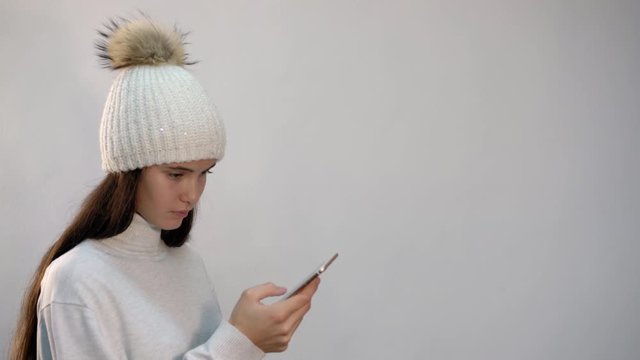 pretty teenage girl in white knitted hat with fur pompom texts in mobile phone on light background close view