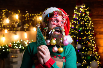 Surprised Santa with New Year balls in beard holds fir-tree. Bearded Santa Claus in party glasses holds small Christmas tree. Christmas beard style. Christmas tree. Merry Christmas and happy new year.