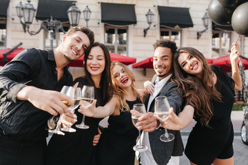 Romantic black-haired woman with bright make-up looking down with smile while having fun with friends outdoor. African man celebrating successful deal and drinking champagne with colleagues.