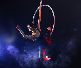 Young woman performing acrobatic element on aerial ring against dark background