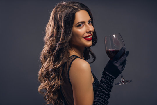 Glamour caucasian brunette woman in evening black dress on a neutral background. She pose sensually with glass of wine. Retro fashion mood.