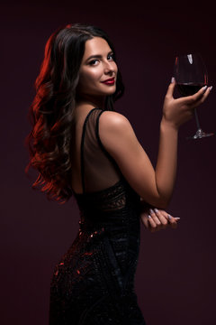 Glamour caucasian brunette woman in evening black dress on a neutral background. She pose sensually with glass of wine. Retro fashion mood.