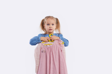 Little child girl chooses outfits for the holiday. Holds hangers with dresses in his hands. White studio background