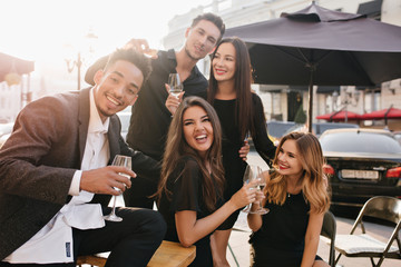 Excited african man with trendy hairstyle says toast while his friends drinking champagne. Outdoor portrait of happy young people celebrating something it warm summer evening.