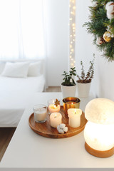 Fototapeta na wymiar Christmas cozy winter home decor. New year interior decorations. White stylish bedroom. Wooden tray with burning candles, night salt lamp on table, bed with white linen, glowing garland lights.