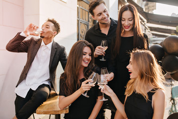 Laughing girl with romantic hairstyle celebrating birthday in outdoor cafe. Blissful friends have fun in sunny day, enjoying weekend together and drinking wine.
