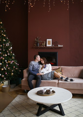 A loving happy couple in cozy warm home clothes hug, kiss and together celebrate Christmas on the background of a decorated Christmas tree, gifts and a fireplace in a cozy house. Girl and guy in love