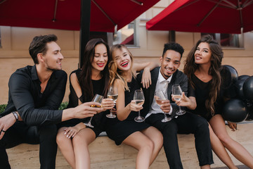 Wonderful blonde girl with red lipstick touching friend's shoulder and laughing in cafe. Portrait of company of young people resting in outdoor cozy restaurant and clink glasses.