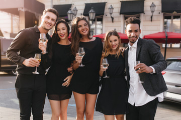 Smiling tanned woman with glass of wine gently embracing blonde sister while posing in front of outdoor cafe. Portrait of ecstatic friends standing on the street after celebration and laughing.