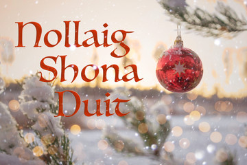 "Nollaig Shona Duit" means "Merry Christmas" in Irish. Blurred winter background of decorated Christmas tree in snow with big red ball. Bokeh. Celebration 25 December. 2020. Greeting card Ireland.