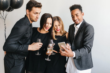 Glad african man showing friends last photos on his phone during party. Handsome man in black attire looking with surprised face expression at smartphone's screen, holding glass of wine.