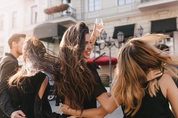 Portrait from back of joyful girls walking around town in windy day and smiling. Brunette woman with long hair waving raising wineglass and looking away, embracing friends.
