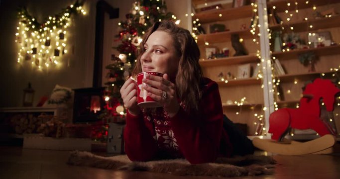 Happy beautiful young girl lies with hot beverage in a cozy s country house by the fireplace and Christmas tree. Dreams and makes a wish