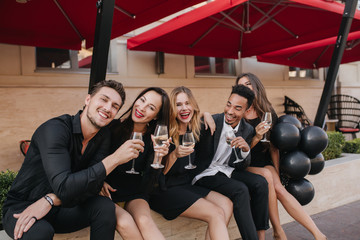 Blonde girl with red lips laughing, while posing between best friends in outdoor cafe. Students in formal attires celebrating graduation and drinking champagne, sitting in open-air restaurant.