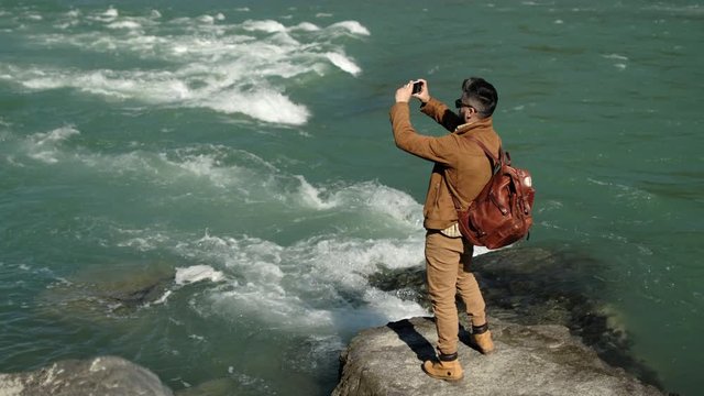 young hiker tourist traveler man taking pictures on smartphone mobile phone by traveling near mountain river. hiking enjoying vacation travel tourism adventure summer landscape nature 4 K slow-mo