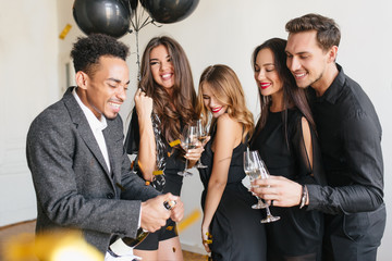 African man in formal attire opens champagne bottle with funny face expression. Refined curly fair-haired lady with trendy make-up holding wineglass during party with best friends.
