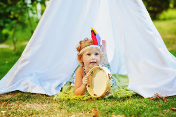 3 years old girl lay on ground near white tent with tambourine in hands and feathers on head. Child pretend to be indian. Indians game outdoors in summertime. Ecological lifestyle for children