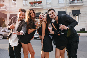 Fototapeta premium Outdoor portrait of laughing students celebrating graduation early in the evening. Ecstatic brunette girl posing with kissing face expression, standing between guy with champagne and woman in dress.