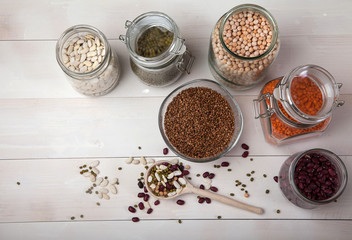Red and white beans, peas, mung bean, red lentils, buckwheat in glass jars and a wooden spoon on a white wooden table. Top view.