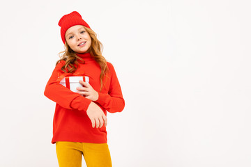 Happy teenager girl with red hair, red hat, hoody and yellow trousers smiles and holds a gift isolated on white background