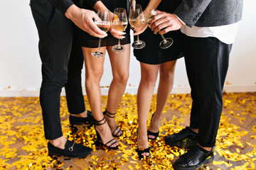 Obraz na płótnie Canvas Indoor portrait of two girls in elegant shoes celebrating new year with boyfriends. Photo of guys in black pants clink wineglasses with ladies on the floor covering with confetti.
