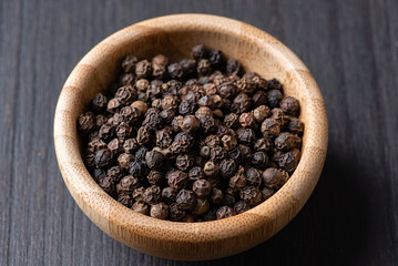 Whole black pepper in a small wooden bowl on a black wooden background. Macro, flat lay.
