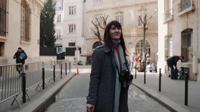 Cheerful beautiful traveler woman with camera posing for camera walking along autumn Paris streets smiling slow motion.