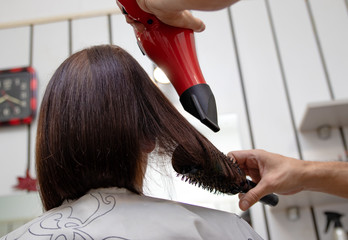 Naklejka premium Hairdresser drying hair and making a hairstyle, using hair brush and hairdryer, to the young brunette woman at the hairdresser salon. Advertising and haircare concept.
