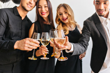 Blur portrait of cute curly woman and her friends with wineglasses on foreground. Fair-haired european female model celebrating new year with colleagues in office.