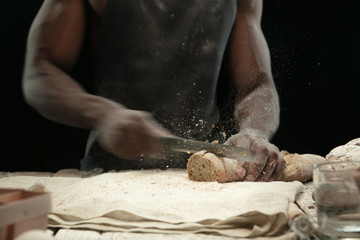 Close up of african-american man slices fresh cereal, white bread, bran with a kitchen knife on wooden table. Healthy eating, nutrition, craft product. Gluten-free food, vegan lifestyle, organic taste
