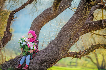 Girl in the forest in a warm coat and a bouquet of flowers in her hands sitting under a tree.