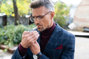Handsome businessman in eyeglasses lighting cigarette on urban street