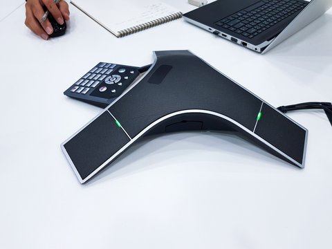 Business conference phone set up in the meeting room. Communication technology for voice conference meeting for long distance office or workplace. With hand taking note on paper, computer laptop.