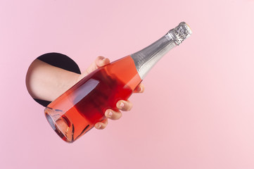 Hand holds a bottle of rose champagne wine through a hole in paper pink background.
