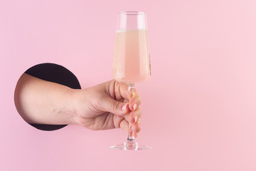 Hand holds a glass of champagne through a hole in paper pink background.