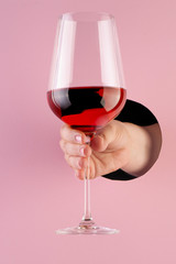 Hand holds a glass of rose wine through a hole in paper pink background.