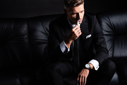 KYIV, UKRAINE - OCTOBER 11, 2019: Businessman smoking iqos while sitting on couch isolated ob black