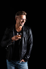Smiling handsome man in leather jacket holding glass of whiskey isolated on black
