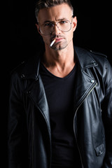 Obraz premium Stylish man in leather jacket smoking cigarette and looking at camera isolated on black