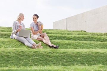 Full length of young businesswomen with disposable coffee cup and laptop sitting on grass steps against sky