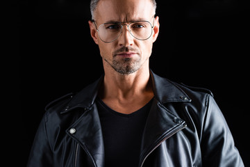 Fototapeta premium Confident stylish man in sunglasses and leather jacket looking at camera isolated on black