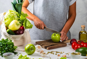 Woman cuts a green apple and cooking a healthy diet food with a cotton eco bag full of fresh vegetables on the table.