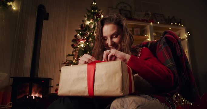 A happy girl hurriedly opens her Christmas gift in a cozy decorated country house near the Christmas tree. Simple moment of happiness