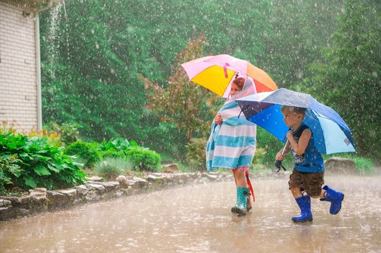 Two Little Kids With Umbrellas And In Rubber Boots Playing Outside During The Rain